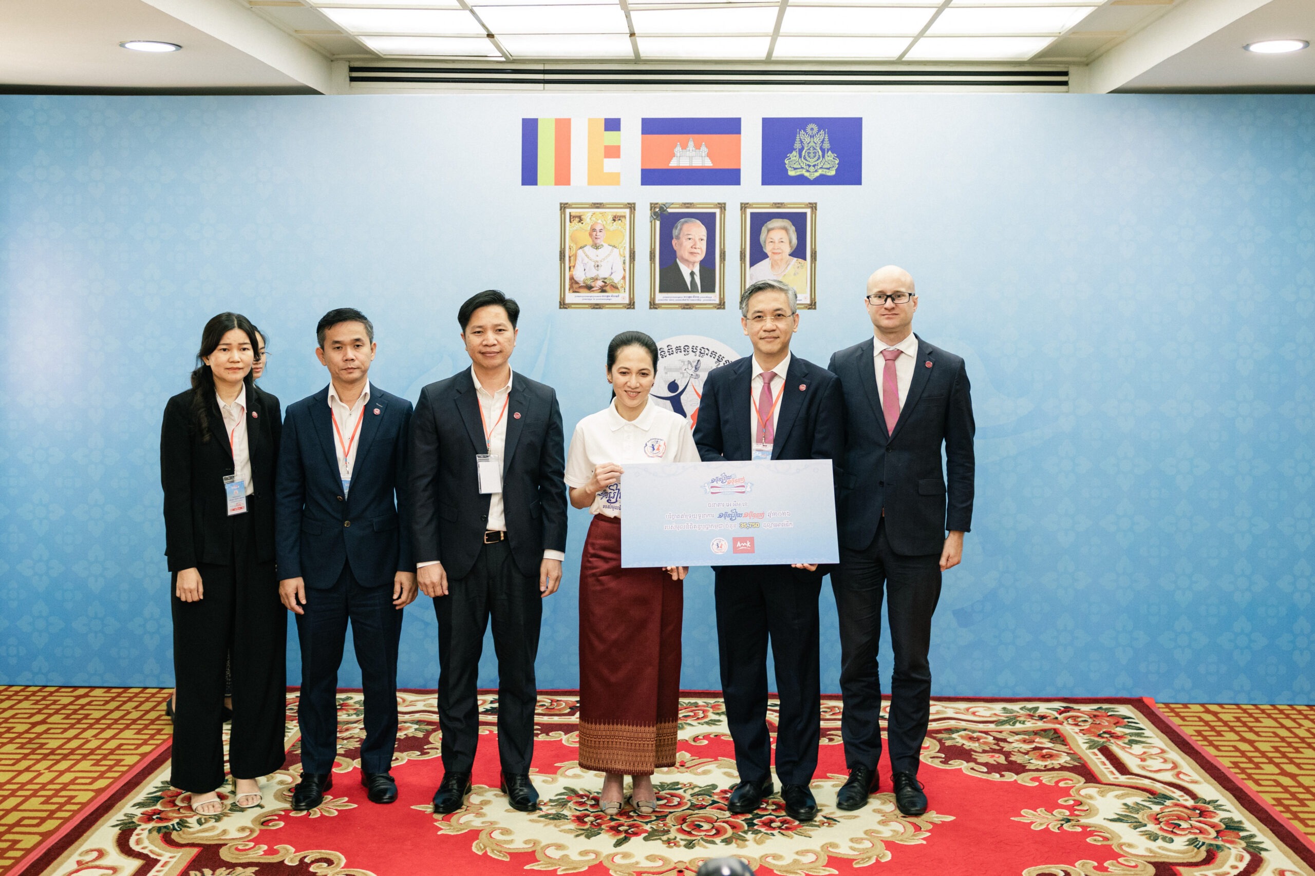 Six professionals in business attire pose with a large ceremonial check in front of a blue backdrop featuring portraits and flags, at a formal event.