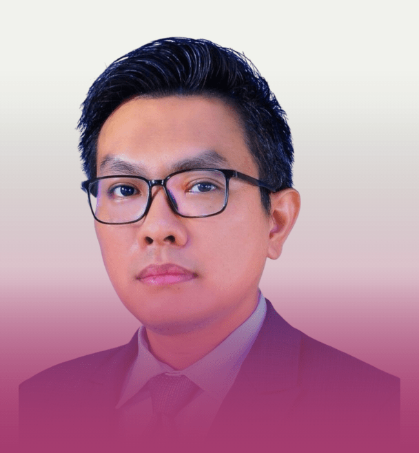 Professional headshot of an Asian man with short black hair and rectangular glasses, wearing a dark suit and tie, looking at the camera with a neutral expression against a white-to-pink gradient background.