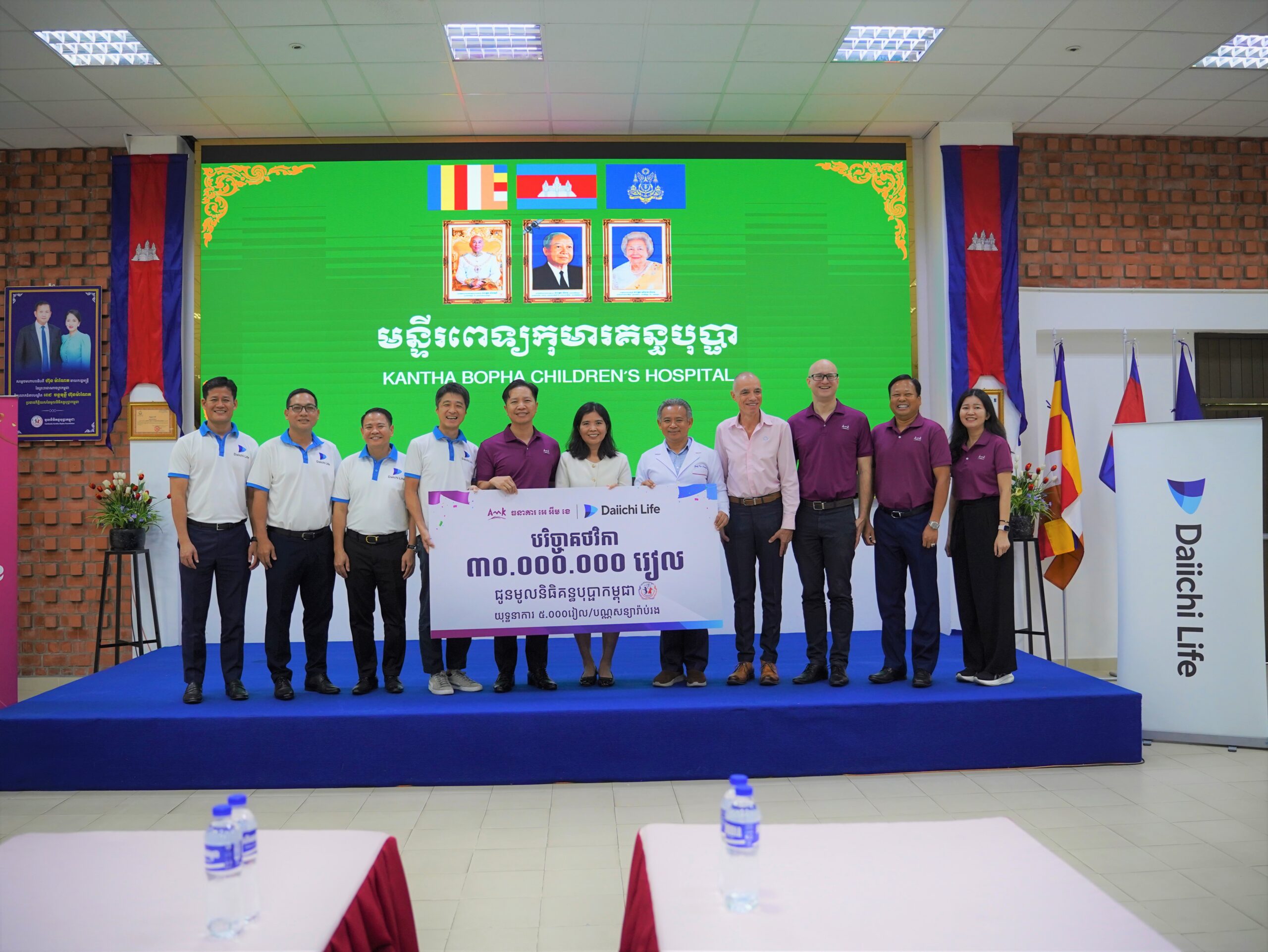 Group of diverse adults standing on a blue stage in front of a green banner reading 'Kantha Bopha Children's Hospital' with portraits above and flags on the sides, celebrating with a large donation banner.